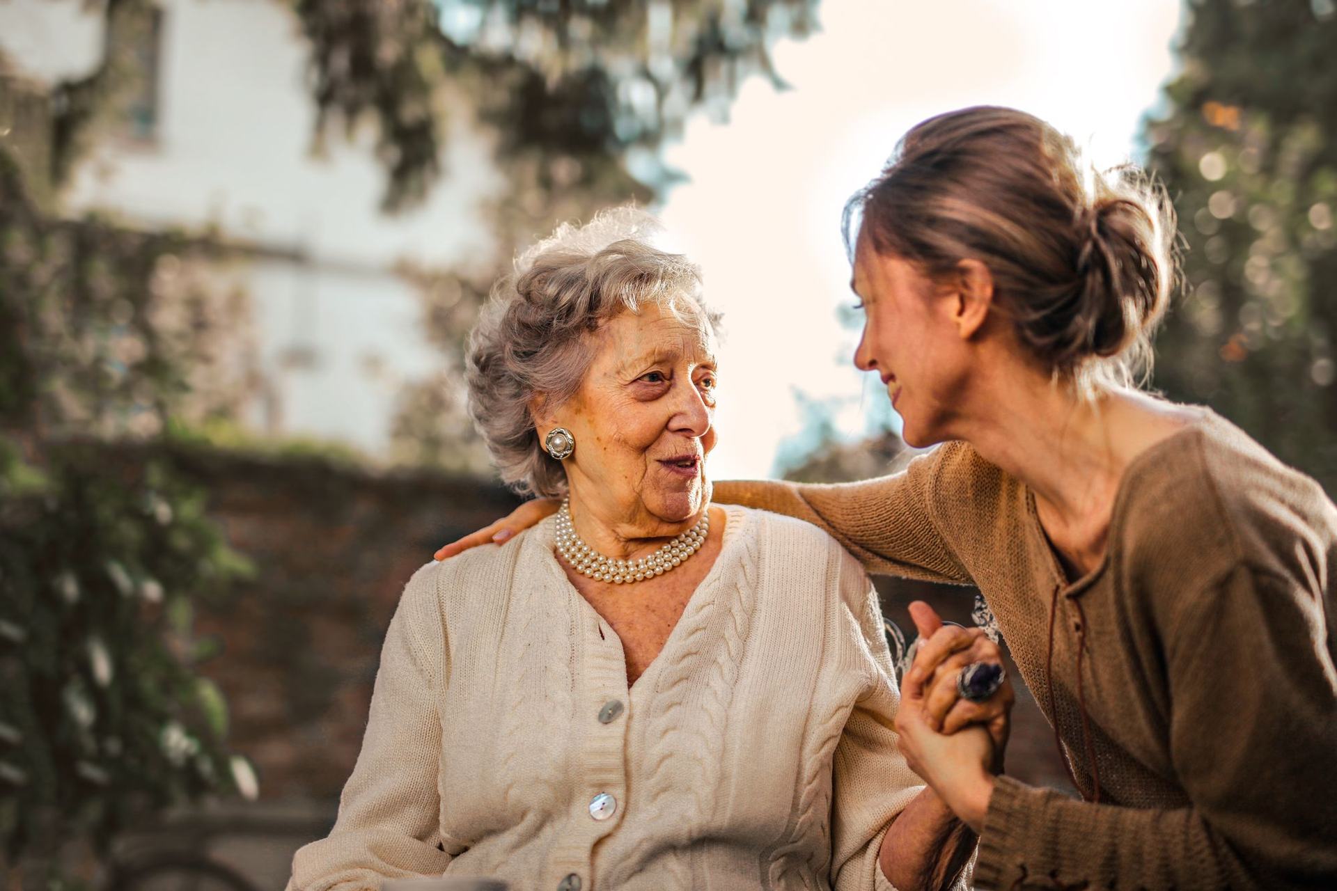 Una hija conversa sonriendo con su madre mayor en un jardín iluminado por el sol